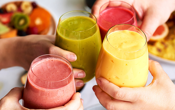 Quatre verres de smoothies colorés levés pour un toast, accompagnés de fruits frais et d’autres plats de brunch en arrière-plan.