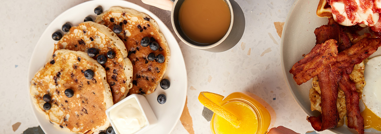 Table garnie d’un brunch varié avec des pancakes aux myrtilles, une omelette aux légumes, du bacon croustillant, des toasts et un jus d’orange frais.