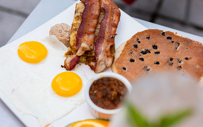 Assiette de brunch comprenant des œufs au plat, du bacon croustillant, un pancake aux myrtilles, des haricots mijotés et une tranche d’orange.