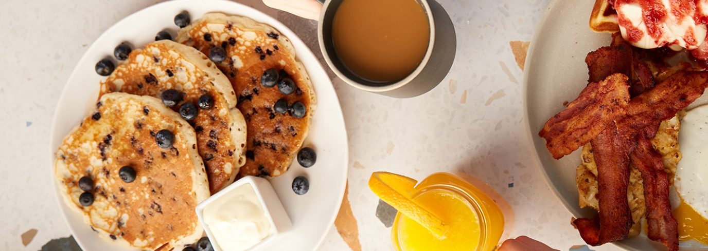 entete Table garnie d’un brunch varié avec des pancakes aux myrtilles, une omelette aux légumes, du bacon croustillant, des toasts et un jus d’orange frais.